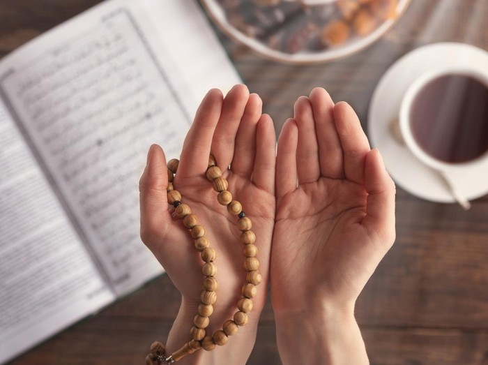 female hand of prayer with wooden beads in sunlight, iftar concept, Ramadan month, Koran, plate of dried fruit, Cup of tea on wooden table