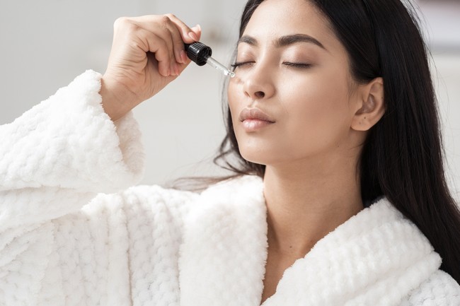 Skincare and beauty concept. Portrait shot of young asian woman applying serum or essential oil on facial skin. Model in white bathrobe moisturizing derma with vitamin E, collagen and hyaluronic acid
