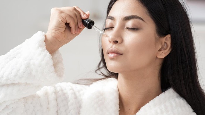 Skincare and beauty concept. Portrait shot of young asian woman applying serum or essential oil on facial skin. Model in white bathrobe moisturizing derma with vitamin E, collagen and hyaluronic acid