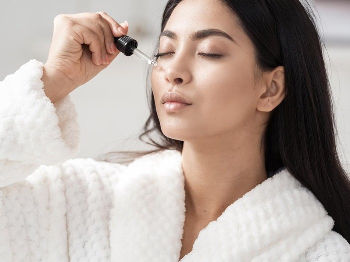 Skincare and beauty concept. Portrait shot of young asian woman applying serum or essential oil on facial skin. Model in white bathrobe moisturizing derma with vitamin E, collagen and hyaluronic acid