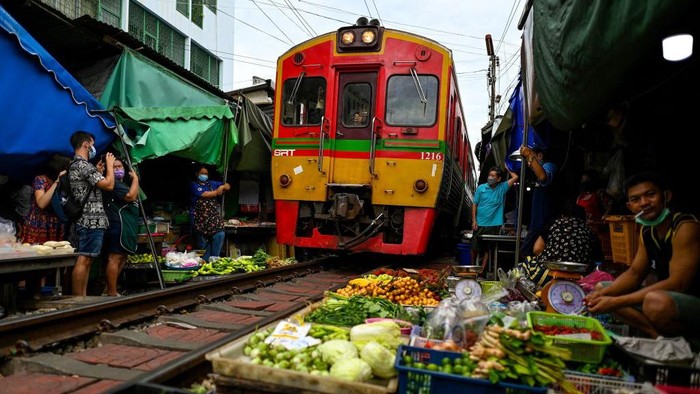 Sensasi Belanja di Pasar Mepet Rel Kereta di Thailand TOPSHOT - This photograph taken on June 4, 2022 shows a passenger train passing through the Mae Klong railway market in Samut Songkhram province, around 80 kms (50 miles) southwest of Bangkok. - Six times a day at the market, local customers and foreign tourists scramble into nooks and crannies while vendors calmly move their woven baskets of goods away from the tracks and close their umbrellas. - TO GO WITH Thailand-tourism-railway-market,FOCUS by Lisa MARTIN (Photo by Manan VATSYAYANA / AFP) / TO GO WITH Thailand-tourism-railway-market,FOCUS by Lisa MARTIN (Photo by MANAN VATSYAYANA/AFP via Getty Images)
