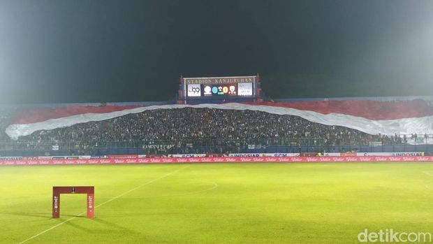 Ada yang berbeda di Stadion Kanjuruhan malam ini. Jelang laga Arema FC vs PSM Makassar, Aremania membentangkan Bendera Merah Putih raksasa di tribun.