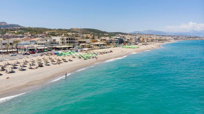 1241286583 Aerial images from a drone of the coast of Rethymno town with the long beach and the beach bars in Creta island. People are seen enjoying the sun under the umbrella at the beach bar or swimming in the crystal clear sea. Rethymno is a historic Mediterranean beach town on the northern coast of Crete, laying on the Aegean Sea with a population of 40.000 people. A touristic destination with a historic Venetian port and town, Archaeological sites, endless sandy beaches, watersports, nice beautiful traditional taverns, and a big variety of hotels. Tourism is bouncing back with reservations exceeding the pre-pandemic numbers, putting Greece and the Greek islands on high demand. Tourists travel after the Covid-19 Coronavirus pandemic. Rethimno, Crete island, Greece on June 13, 2022 (Photo by Nicolas Economou/NurPhoto via Getty Images)