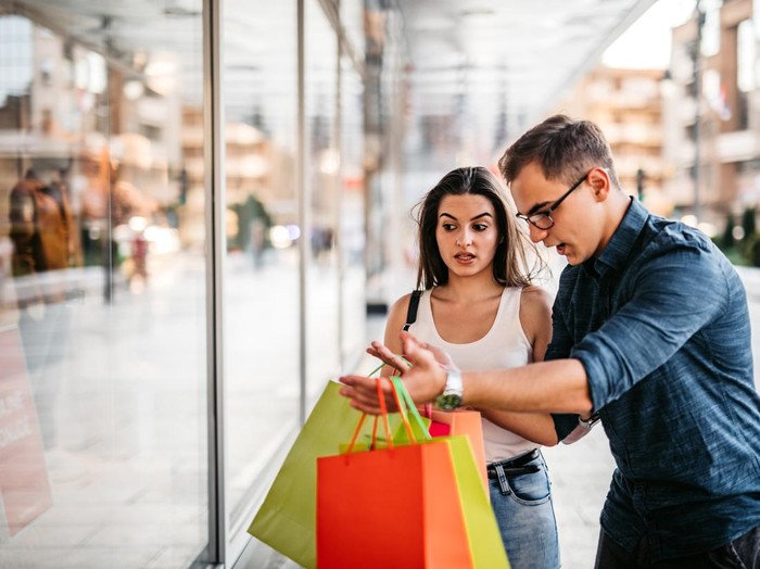 1280385738 Young man scolding his girlfriend in front of the shop window for spending money on shopping, carrying in his hands a bunch of shopping bags.