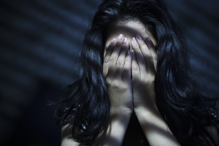 Indoor image of a young woman covering her face with her both hands out of embracement and sadness. Striped Light and shadows are falling on her through blinds. One person, low key, horizontal composition with selective focus and copy space.