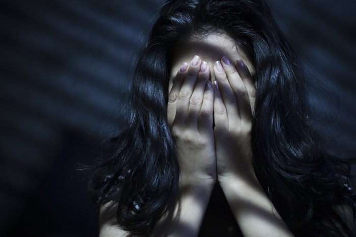 612508910 Indoor image of a young woman covering her face with her both hands out of embracement and sadness. Striped Light and shadows are falling on her through blinds. One person, low key, horizontal composition with selective focus and copy space.