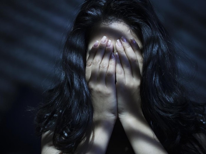 612508910 Indoor image of a young woman covering her face with her both hands out of embracement and sadness. Striped Light and shadows are falling on her through blinds. One person, low key, horizontal composition with selective focus and copy space.