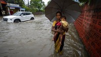 Seorang wanita menggendong seorang anak dan mengarungi jalan yang tergenang banjir. India di bagian timur mengalami volume air hujan yang tinggi. Sementara India di sisi lain seperti di Delhi sedang mengalami cuaca kering dan panas.