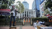 Salah satu peserta aksi berorasi, sementara dua orang peserta yang menggambarkan pasangan suami istri tampak membaca dokumen di atas kasur.