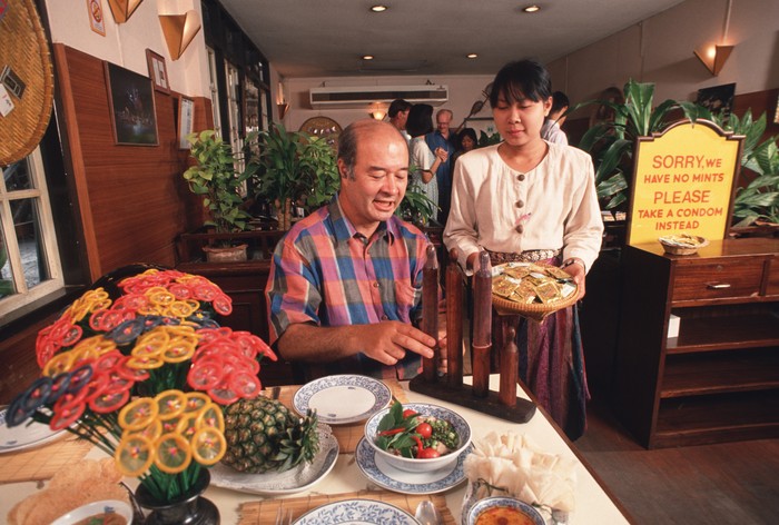 158608233 BANGKOK, THAILAND - 1994/02/01: A portrait of Mechai Viravaidya who is Thailand's leading anti AIDS activist sitting in the restaurant he founded called 'Cabbages and Condoms. Here he takes an 'after-dinner condom' instead of a mint! He has popularised condom use in Thailand and has created popular family planning clinics in the country. Such is his fame that condoms in Thailand are now known as 'Mechais'.. (Photo by Peter Charlesworth/LightRocket via Getty Images)