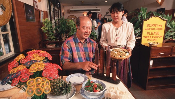 158608233 BANGKOK, THAILAND - 1994/02/01: A portrait of Mechai Viravaidya who is Thailands leading anti AIDS activist sitting in the restaurant he founded called Cabbages and Condoms. Here he takes an after-dinner condom instead of a mint! He has popularised condom use in Thailand and has created popular family planning clinics in the country. Such is his fame that condoms in Thailand are now known as Mechais.. (Photo by Peter Charlesworth/LightRocket via Getty Images)