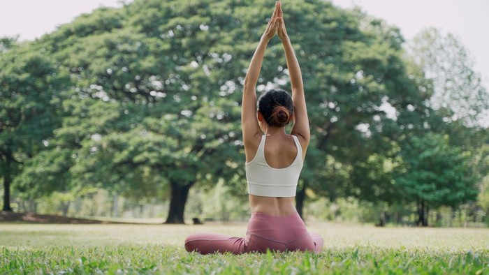 Hari Yoga Internasional 2022 Back view of an unrecognizable slender Asian young woman sitting on the grass in the lotus position and raising hands up outside in city park with the big trees background. Rearview of female practicing yoga outdoors on a sunny day.