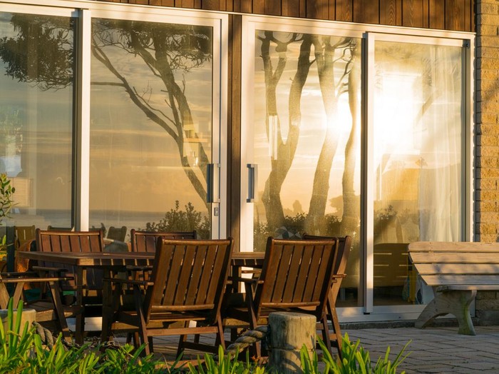 Waterfront home exterior with outdoor furniture and large windows reflecting sunrise over the sea