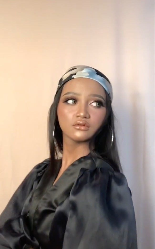 Tanpa merubah warna kulitnya, gadis tersebut didandani dengan makeup glowing bernuansa nude dan softlens berwarna terang. Sambil mengenakan dress hitam, ikat kepala, dan anting bulat yang cukup besar, penampilan sang gadis pun tampak mempesona. Foto: TikTok/@panggilakunano
