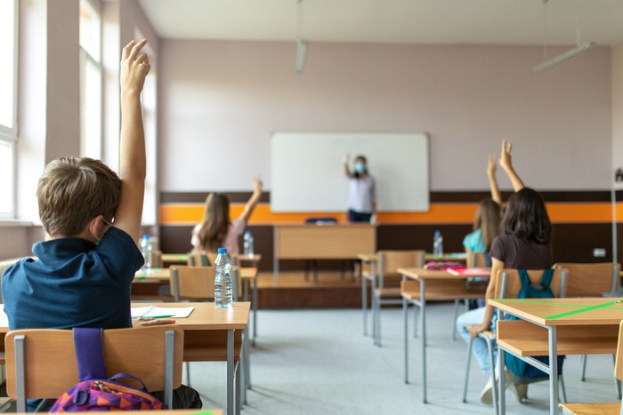 Students with protective masks sitting in school desks in their classroom. School desks are marked with a cross to mark a place where sitting is not allowed to maintain social distance during corona virus .