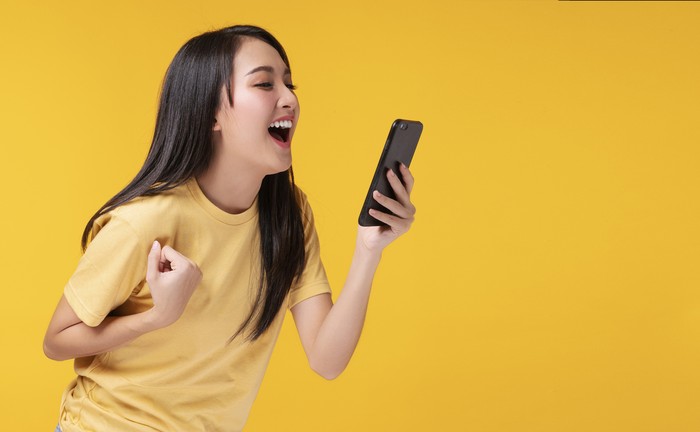 Cheerful young woman holding smartphone and her hand with shocked amazed for success or get good news over isolated yellow background.