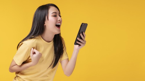 Cheerful young woman holding smartphone and her hand with shocked amazed for success or get good news over isolated yellow background.