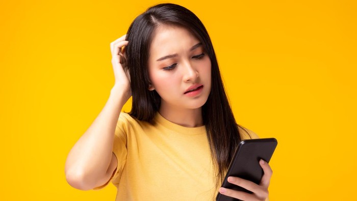 Beautiful young woman get confused and frustrated of application shopping online Girl hold mobile phone, look at cellphone with expression face She get disappointed of broken phone yellow background