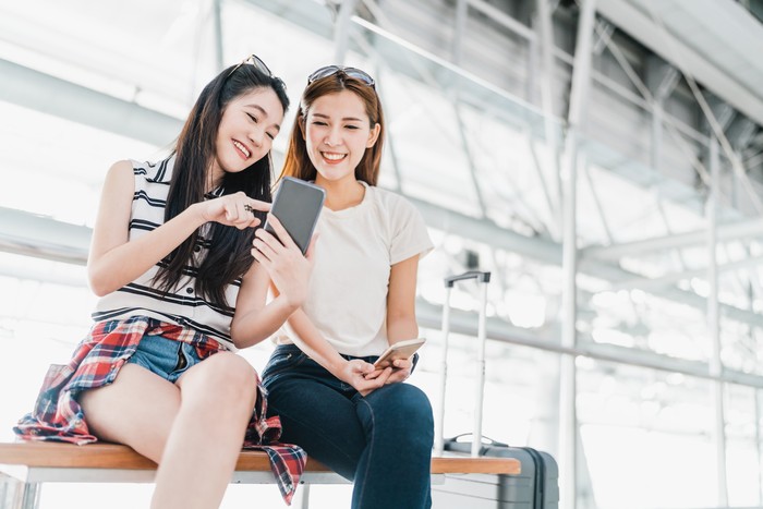 Two happy Asian girls using smartphone checking flight or online check-in at airport together, with luggage. Air travel, summer holiday, or mobile phone application technology concept