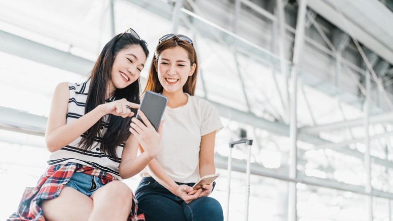 Ilustrasi Turis China Two happy Asian girls using smartphone checking flight or online check-in at airport together, with luggage. Air travel, summer holiday, or mobile phone application technology concept