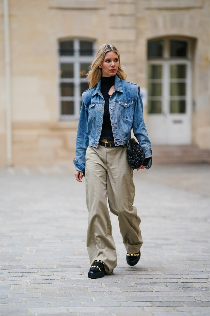 PARIS, FRANCE - APRIL 05: Natalia Verza @mascarada.paris wears a black turtleneck / cut-out asymmetric shoulder / long sleeves body, a blue denim oversized jacket, a black shiny leather with gold small YSL buckle belt from Saint Laurent Paris, a black shiny braided leather puffy handbag, beige large flared pants, black shin leather with embroidered gold metallic pearls strappy / platform soles sandals, black socks, during a street style fashion photo session, on April 05, 2022 in Paris, France. (Photo by Edward Berthelot/Getty Images)