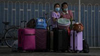 Bergeser ke terminal bus, para wanita mengenakan masker menunggu bus di samping koper mereka. (AP News)