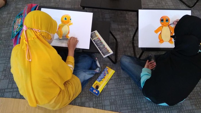 Sulis dan Nayla, peserta Bootcamp Kelas Gambar dan Handai Tuli menggambar boneka Charizard dengan medium krayon di atas kertas gambar.