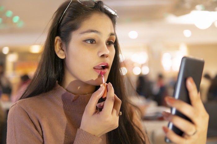 Indoor image of an Asian, Indian beautiful young woman wearing eyeglasses head, sitting at a restaurant and applying lipstick while looking at the phone screen.