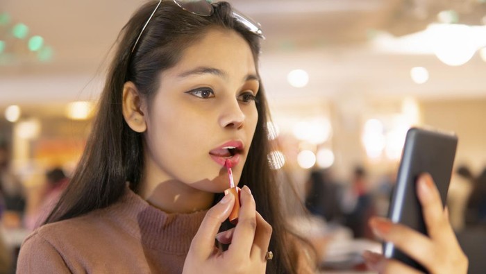 Indoor image of an Asian, Indian beautiful young woman wearing eyeglasses head, sitting at a restaurant and applying lipstick while looking at the phone screen.