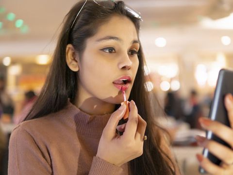 Indoor image of an Asian, Indian beautiful young woman wearing eyeglasses head, sitting at a restaurant and applying lipstick while looking at the phone screen.