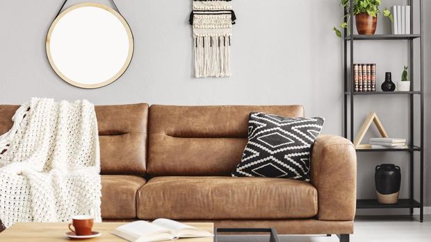 Real photo of handmade macrame and mockup poster hanging on the wall in bright sitting room interior with leather sofa and metal rack with decor and books