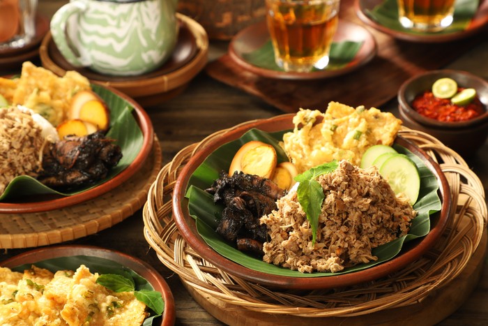Nasi Megono, traditional Javanese rice dish from Pekalongan, Central Java. It is steamed rice with Megono (stir-fried minced young jackfruit) as the main side dish; then added with Cumi Hitam (braised squid in its ink), Pindang Telur (braised egg in soy sauce) and Tempe Mendoan (fried tempeh in batter). The dish is plated on an earthenware plate lined with banana leaf. Served with Sambal (chili sauce) as the condiment. The Nasi Megono and Sambal are arranged on a table with another portion of Nasi Megono, extra portion of Tempe Mendoan and hot tea with the tea pot.