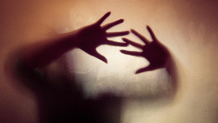 Colour backlit image of the silhouette of a woman with her hands pressed against a glass window. The silhouette is distorted, and the arms elongated, giving an alien-like quality. The image is sinister and foreboding, with an element of horror. It is as if the woman is trying to escape from behind the glass. Horizontal image with copy space.