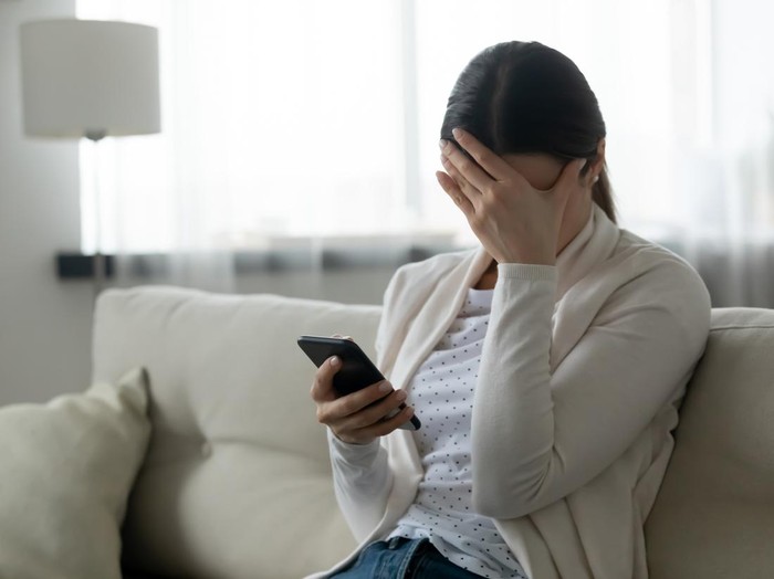 Woman sit on sofa holding smartphone cover face with hand feels scared humiliated suffering from cyberbullying being on-line abused by stalker. Bad news, life troubles, break up with boyfriend concept