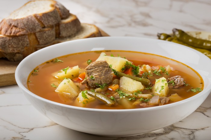 Bowl of vegetable beef soup with sliced bread and hot chilli peppers in background