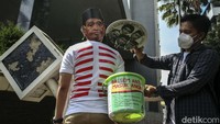 Pria yang memegangi koin itu mulanya tampak melakukan gerakan mengambil balsem dari ember tersebut. Kemudian, dia menggaruk bagian belakang punggung pria bertopeng Tumpak.