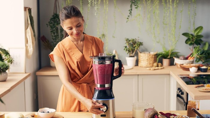 Young female pressing start button of blender during preparation of homemade vegetable smoothie by kitchen table