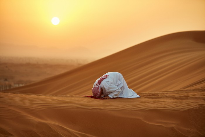 Arab man praying on mat in desert. Male is in traditional wear. He is kneeling on sand.