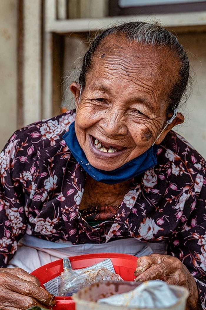 This photo taken on June 22, 2022 shows waiting customers gathering round septuagenarian Mbah Satinem (centre R) as she prepares lupis, a traditional sweet dish made from glutinous rice and served with grated coconut and liquid palm sugar, at her street stall in Yogyakarta. - As the morning twilight approaches in an Indonesian city famed for its street food scene, a queue of sleepy customers snakes its way along the road in anticipation of a local culinary legend. - TO GO WITH AFP STORY INDONESIA-FOOD-DRINK-COOKING,FOCUS BY DESSY SAGITA (Photo by JUNI KRISWANTO / AFP) / TO GO WITH AFP STORY INDONESIA-FOOD-DRINK-COOKING,FOCUS BY DESSY SAGITA (Photo by JUNI KRISWANTO/AFP via Getty Images)