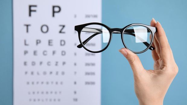 Woman holding glasses against eye chart on blue background, closeup. Ophthalmologist prescription