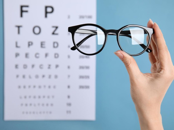 Woman holding glasses against eye chart on blue background, closeup. Ophthalmologist prescription