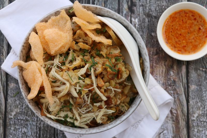 A popular Peranakan breakfast and fast food dish in Indonesia, consists of rice porridge with toppings of shredded chicken, sliced Chinese croissant, fried shallot, chopped celery leaves, and crisp crackers. Accompanied with chili sauce. Typically an inexpensive, street food dish, it is now served in a fancy ceramic bowl with Chinese-style, ceramic soup spoon tucked on the upper right hand side. A casually-folded white linen napkin underlines the bowl. The meal is served on an unpolished, rustic wooden table. The image was taken outdoor in the morning and used sunlight as the natural lighting source.