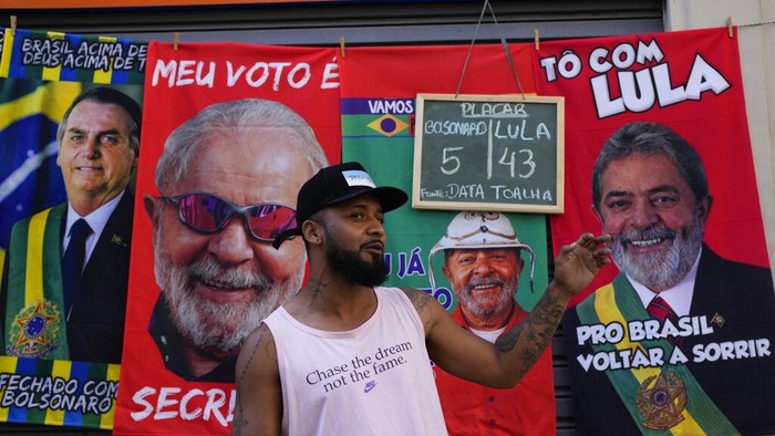 Jose Lucas da Silva stands in front of his towels for sale featuring Brazilian President Jair Bolsonaro, far left, and former President Luiz Inacio da Silva, or Lula, and his chalkboard showing his daily sales count for each presidential contender towel, ahead of elections in Rio de Janeiro, Brazil, Wednesday, July 27, 2022. General elections are set for Oct. 2. (AP Photo/Silvia Izquierdo)