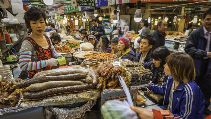 Penduduk setempat duduk dan makan makanan tradisional Korea di sebuah warung makan jalanan yang ramai yang disajikan oleh seorang pedagang wanita di Pasar Dongdaemun yang semarak dan ramai di jantung pusat kota Seoul, Korea.