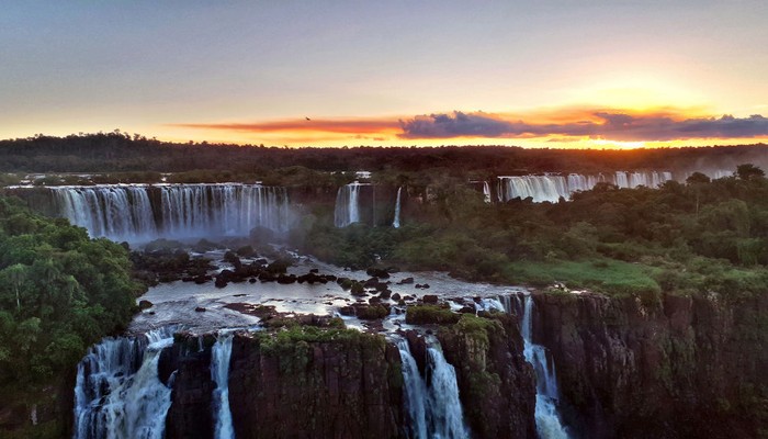 1169311745 IGUACU, BRAZIL - APRIL 8: Tourists enjoy Brazilian Caipirinha cocktails, made with Cachaca, fruit and sugar, in front of the evening panoramic view of the Argentinian side of the waterfalls on April 8, 2019 in the IguaΓ§u National Park, Brazil. The falls, which straddle the border of Argentina and Brazil, are known in Spanish as Cataratas del IguazΓΊ and in Portuguese as Cataratas do IguaΓ§u. They are the largest waterfall system in the world made up of 275 individual drops stretching 2.7 kilometers wide and up to 82 meters in height, and were chosen in 2011 as one of the New Seven Wonders of Nature. The Argentinian and Brazilian national parks which protect the falls host millions of tourists each year while protecting the environment by limiting vehicle access and using ecological transportation systems within the parks. (Photo by David Silverman/Getty Images)