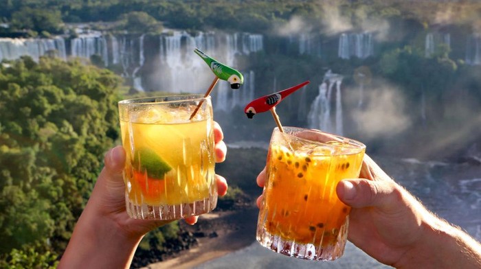 1169311745 IGUACU, BRAZIL - APRIL 8: Tourists enjoy Brazilian Caipirinha cocktails, made with Cachaca, fruit and sugar, in front of the evening panoramic view of the Argentinian side of the waterfalls on April 8, 2019 in the IguaΓ§u National Park, Brazil. The falls, which straddle the border of Argentina and Brazil, are known in Spanish as Cataratas del IguazΓΊ and in Portuguese as Cataratas do IguaΓ§u. They are the largest waterfall system in the world made up of 275 individual drops stretching 2.7 kilometers wide and up to 82 meters in height, and were chosen in 2011 as one of the New Seven Wonders of Nature. The Argentinian and Brazilian national parks which protect the falls host millions of tourists each year while protecting the environment by limiting vehicle access and using ecological transportation systems within the parks. (Photo by David Silverman/Getty Images)
