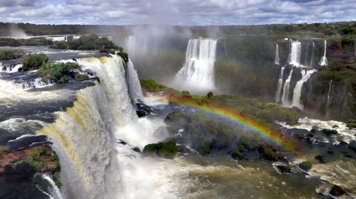 1169311745 IGUACU, BRAZIL - APRIL 8: Tourists enjoy Brazilian Caipirinha cocktails, made with Cachaca, fruit and sugar, in front of the evening panoramic view of the Argentinian side of the waterfalls on April 8, 2019 in the IguaΓ§u National Park, Brazil. The falls, which straddle the border of Argentina and Brazil, are known in Spanish as Cataratas del IguazΓΊ and in Portuguese as Cataratas do IguaΓ§u. They are the largest waterfall system in the world made up of 275 individual drops stretching 2.7 kilometers wide and up to 82 meters in height, and were chosen in 2011 as one of the New Seven Wonders of Nature. The Argentinian and Brazilian national parks which protect the falls host millions of tourists each year while protecting the environment by limiting vehicle access and using ecological transportation systems within the parks. (Photo by David Silverman/Getty Images)