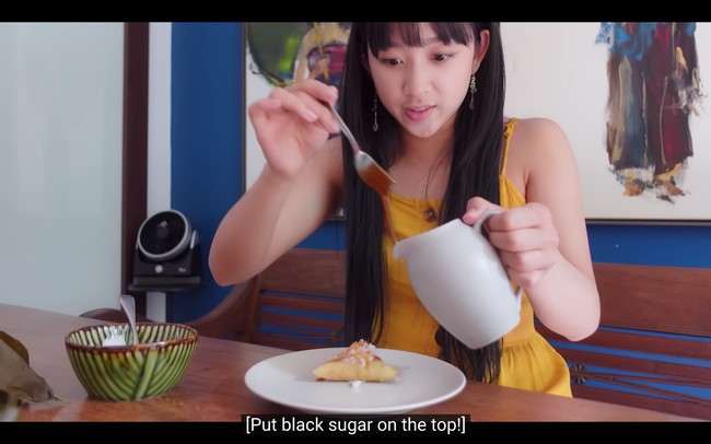 Lewat vlognya, Dita Karang juga memperkenalkan kue lapis sebagai dessert Indonesia kepada penggemar mancanegara. Dia memakai dress berwarna kuning dan anting panjang bertuliskan aksara Jawa. Foto: dok. YouTube Secret Number