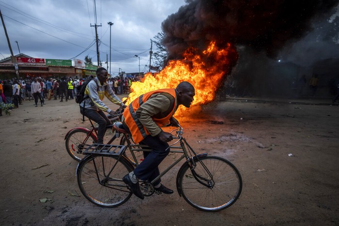Men ride their bicycles past a roadblock of burning tires in the Kibera neighborhood of Nairobi, Kenya Monday, Aug. 15, 2022. After last-minute chaos that could foreshadow a court challenge, Kenya's electoral commission chairman has declared Deputy President William Ruto the winner of the close presidential election over five-time contender Raila Odinga. (AP Photo/Ben Curtis)