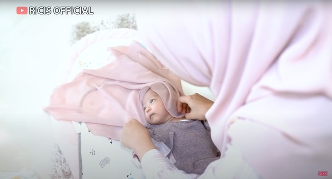 Dengan hati-hati Ricis berusaha memasangkan kerudung di kepala anaknya. Kerudungnya itu tampak berwarna senada dengan baju yang dikenakan oleh baby Moana. Foto: YouTube/Ricis Official